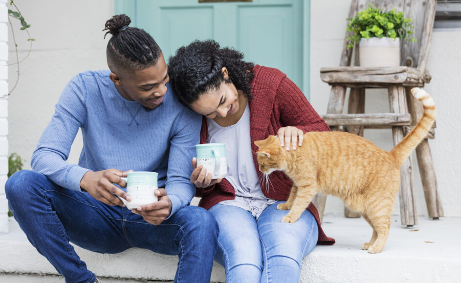 A young African-American couple hanging out together, drinking coffee on the front steps of their home, relaxing and petting a cat.